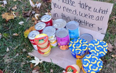 A simple cardboard sign sits behind canned goods that reads Please take what you need, we're all in this together.